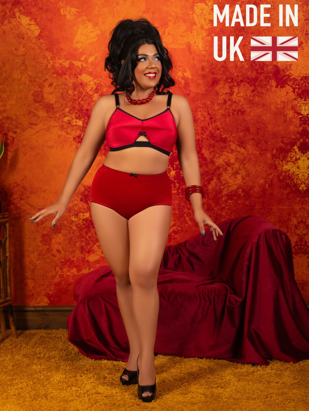 A woman models a two-piece lingerie set consisting of a red bralette with black trim and matching red briefs, standing in front of a textured orange and red studio backdrop with a draped red sofa. A 'MADE IN UK' graphic with a Union Jack flag is in the top right corner.