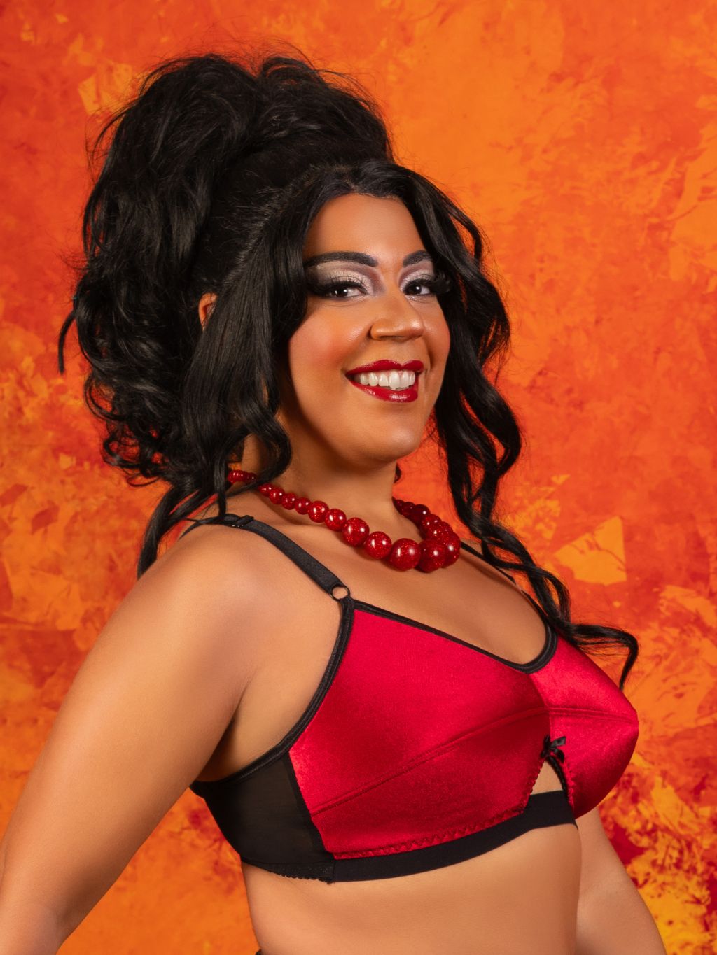 A smiling woman with dark curly hair wearing a red and black bralette and a red beaded necklace, posed against a textured orange background.