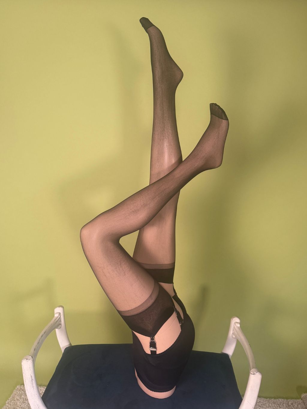 A person wearing sheer black thigh-high stockings and a black garter belt sits upside down on a chair against a yellow-green wall.