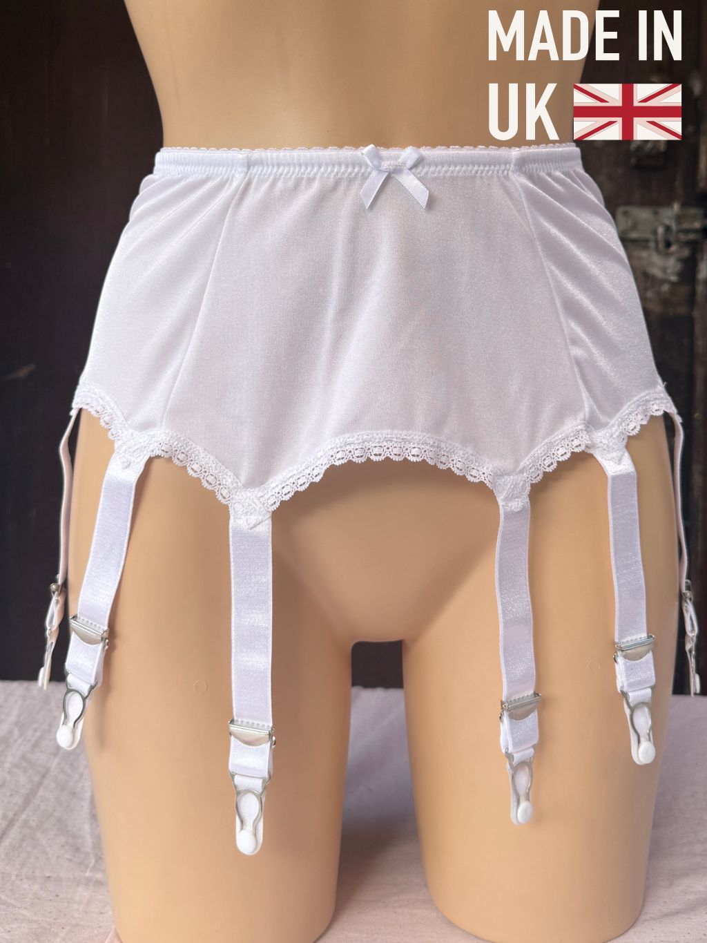White lace-trimmed garter belt with attached suspender straps and metal clips, displayed on a beige mannequin torso. The image includes a 'MADE IN UK' text overlay with a Union Jack flag in the top right corner.