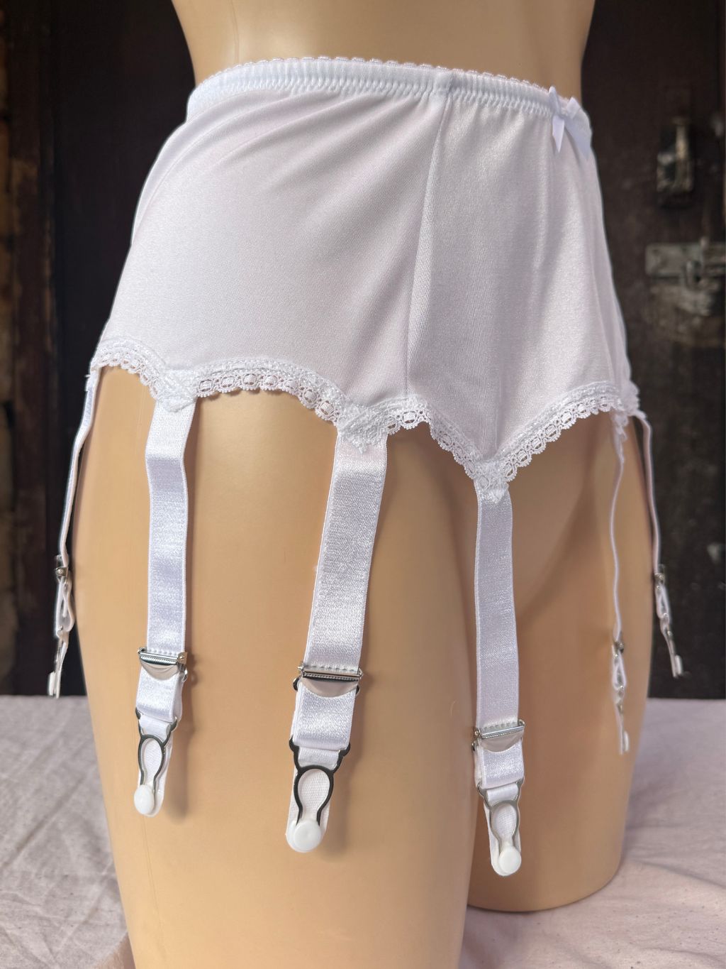 White satin garter belt with lace trim and four attached white garter straps and silver metal clips, displayed on a beige mannequin torso against a dark rustic background.