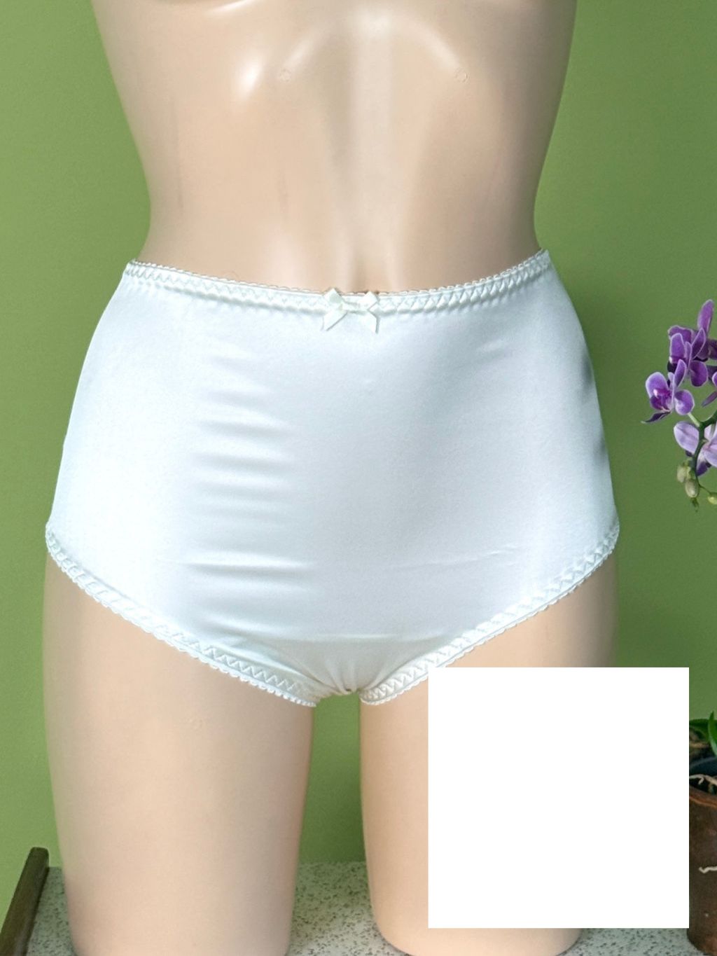White high-waisted briefs displayed on a beige mannequin torso against a green wall, with a small bow at the center front and lace trim at the waistband and leg openings.
